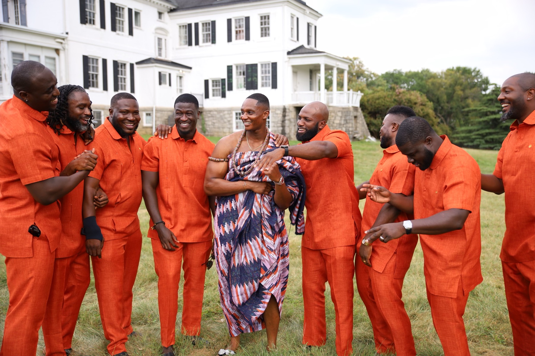 Groom in kente surrounded by his groomsmen in orange, laughing together