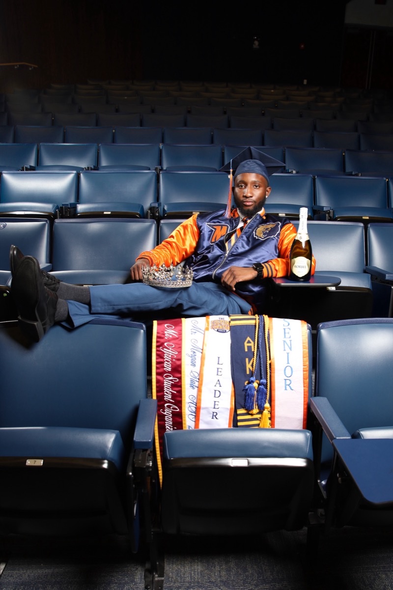 Graduate in cap and gown reclining across auditorium seats with sashes and a crown