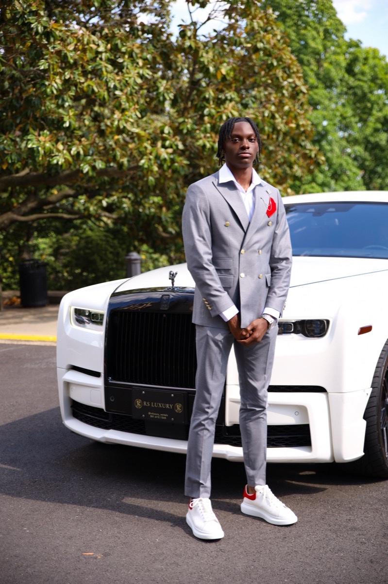 Man in a grey suit posing beside a white luxury car