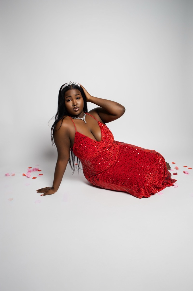 Woman in a red sequin gown and tiara seated on a studio backdrop
