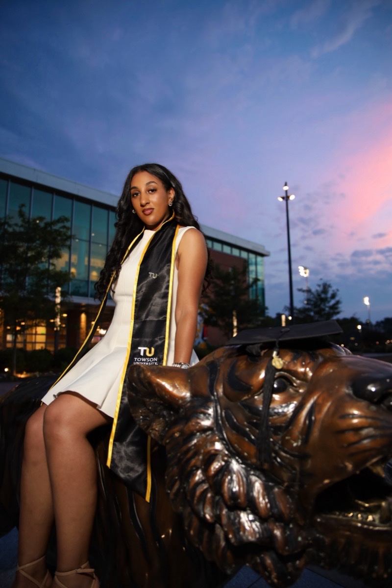 Graduate in cap, gown, and stole posing beside a bronze tiger statue at dusk