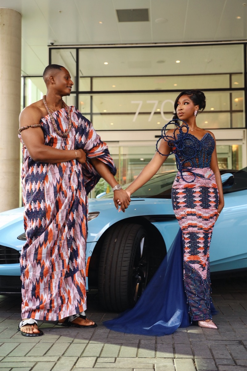 Couple in coordinated kente attire holding hands beside a sports car